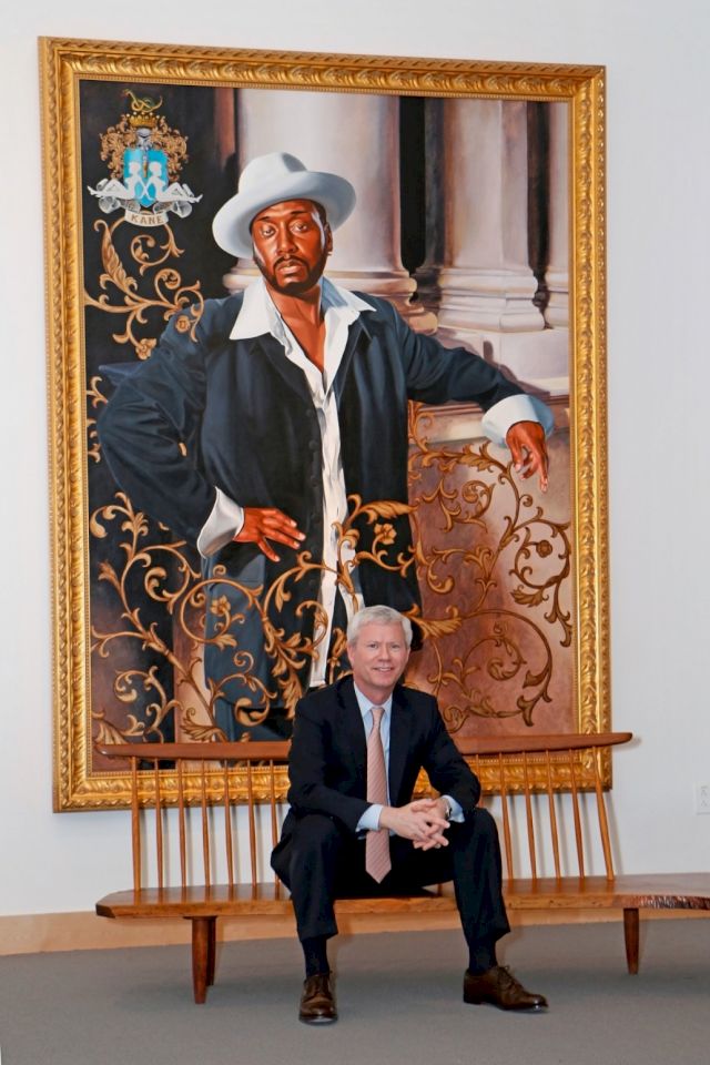John Morrissey seated on a George Nakashima walnut Conoid bench in front of the painting 'Big Daddy Kane' by Kehinde Wiley